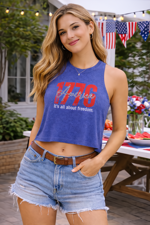 Woman in a blue 1776 America crop top with raw-cut sleeves and denim shorts, outdoors with patriotic decor.