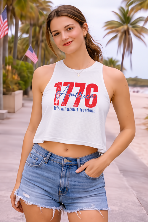 Woman in white 1776 America crop top with raw-cut sleeves, blue denim shorts, and a gold cross necklace.
