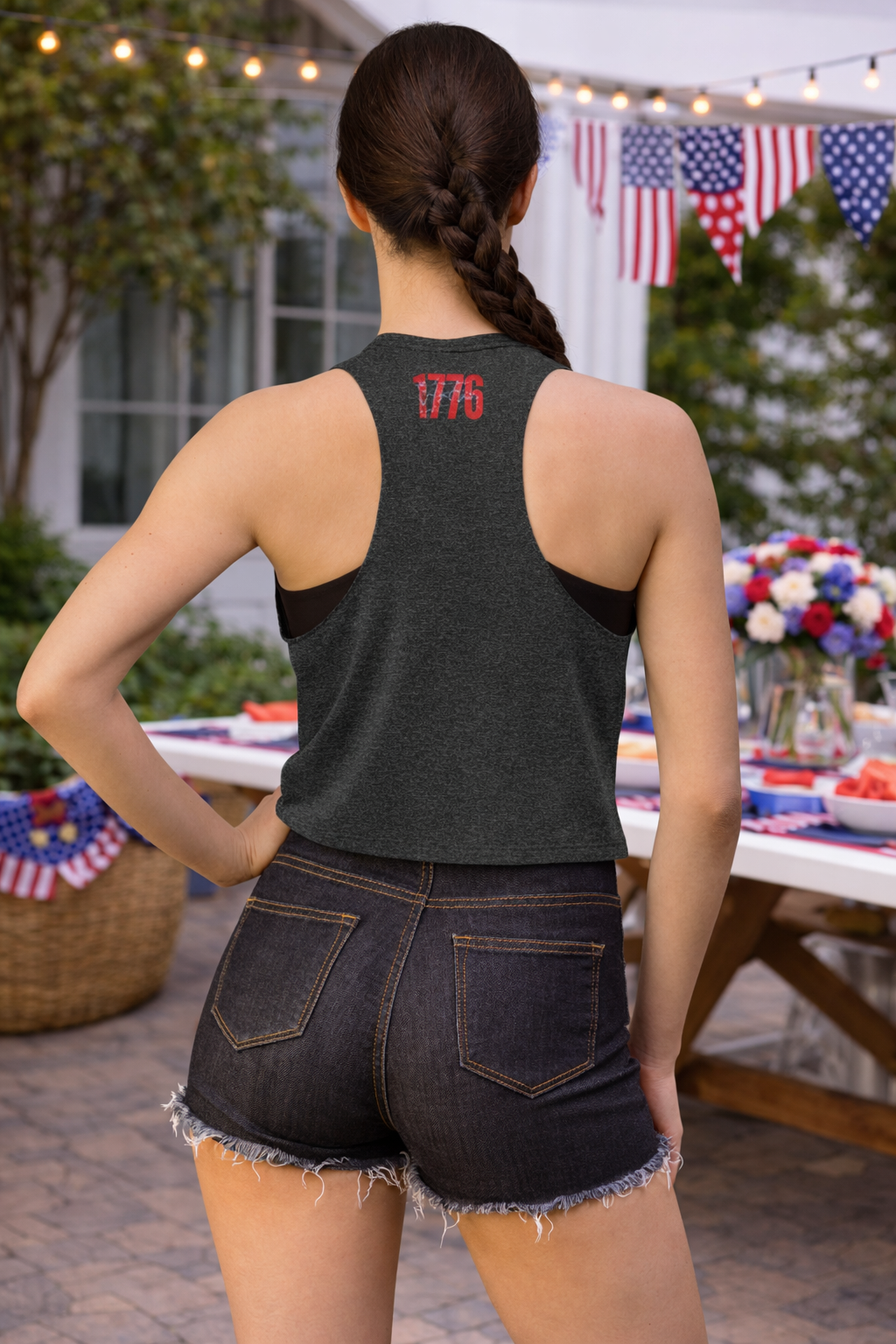Person wearing a tank top with '1776' on the back, standing outdoors with a table and flags in the background.