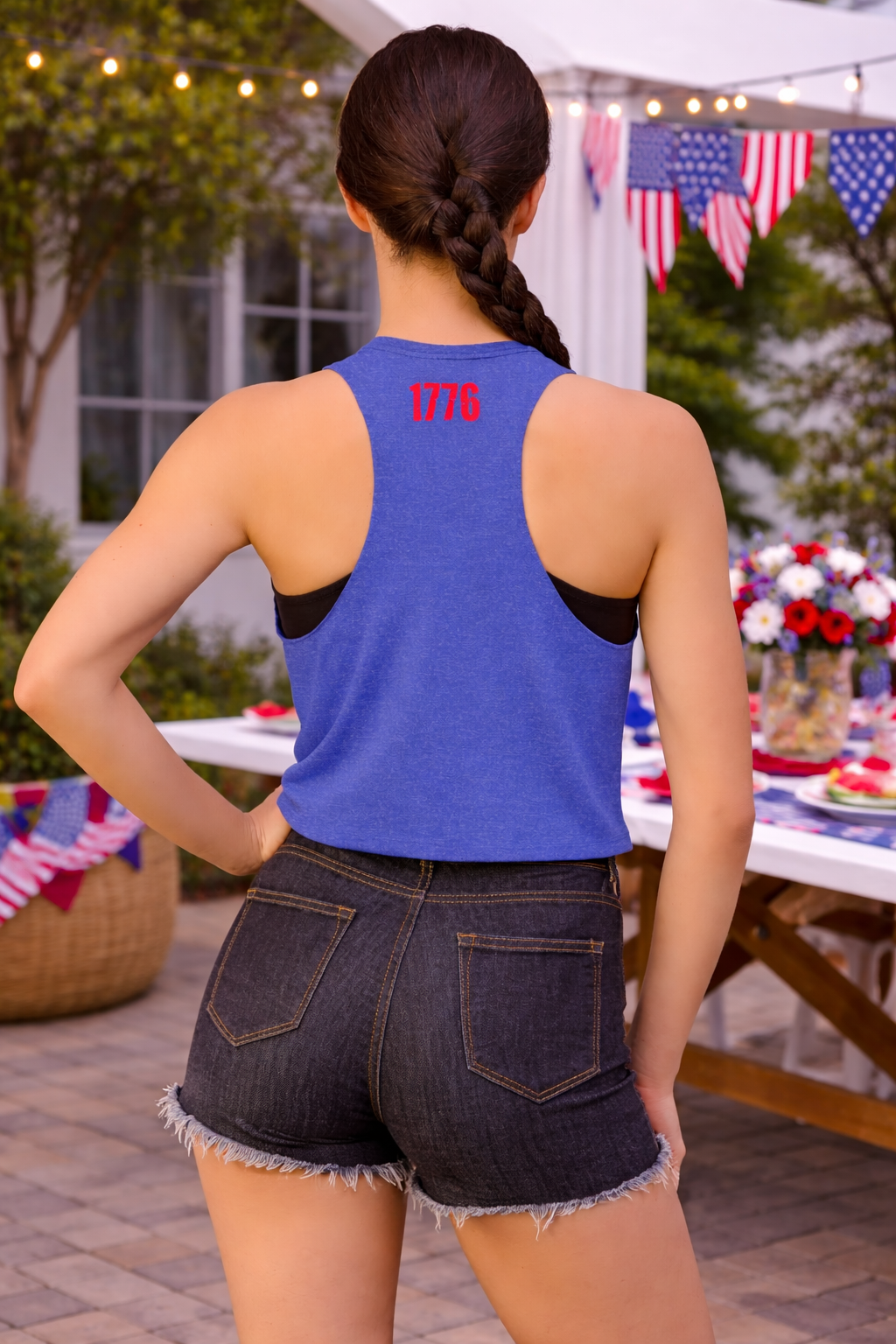 Person wearing a blue tank top with '1776' on the back, standing outdoors with a table and flags in the background.