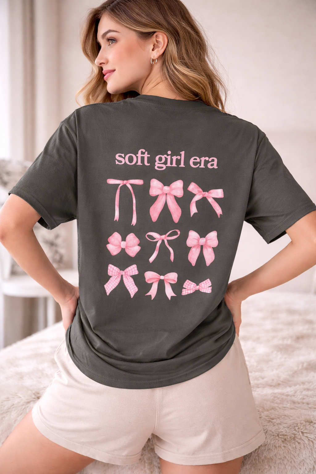 Woman wearing a dark gray t-shirt with 'soft girl era' and pink bows on the back.