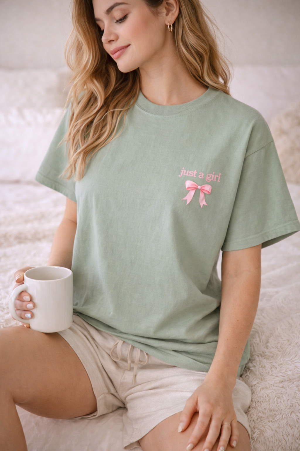 Woman wearing a green t-shirt with a pink bow design, sitting on a bed holding a white mug.