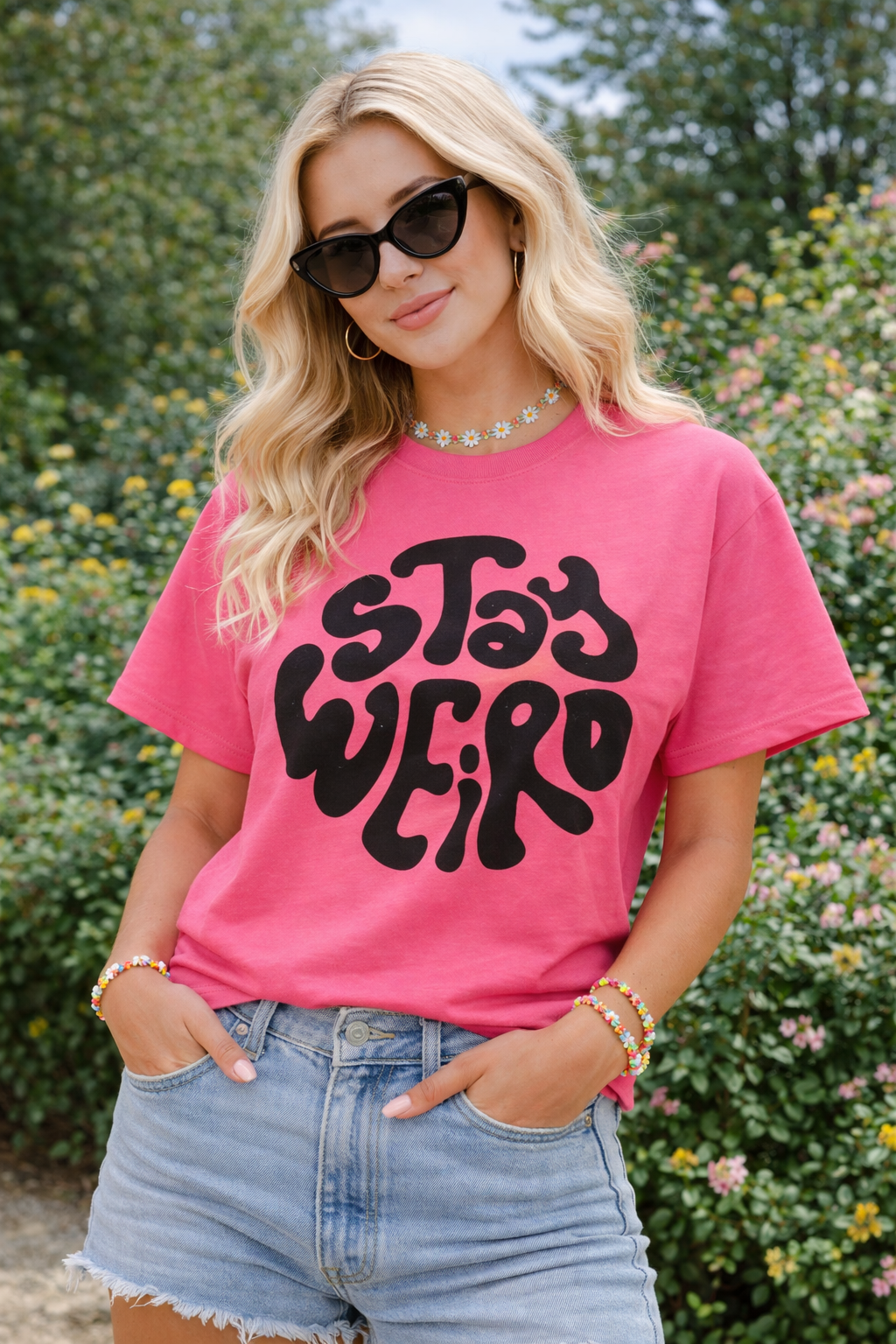 Woman wearing a pink t-shirt with 'Stay Weird' text, sunglasses, and denim shorts outdoors.