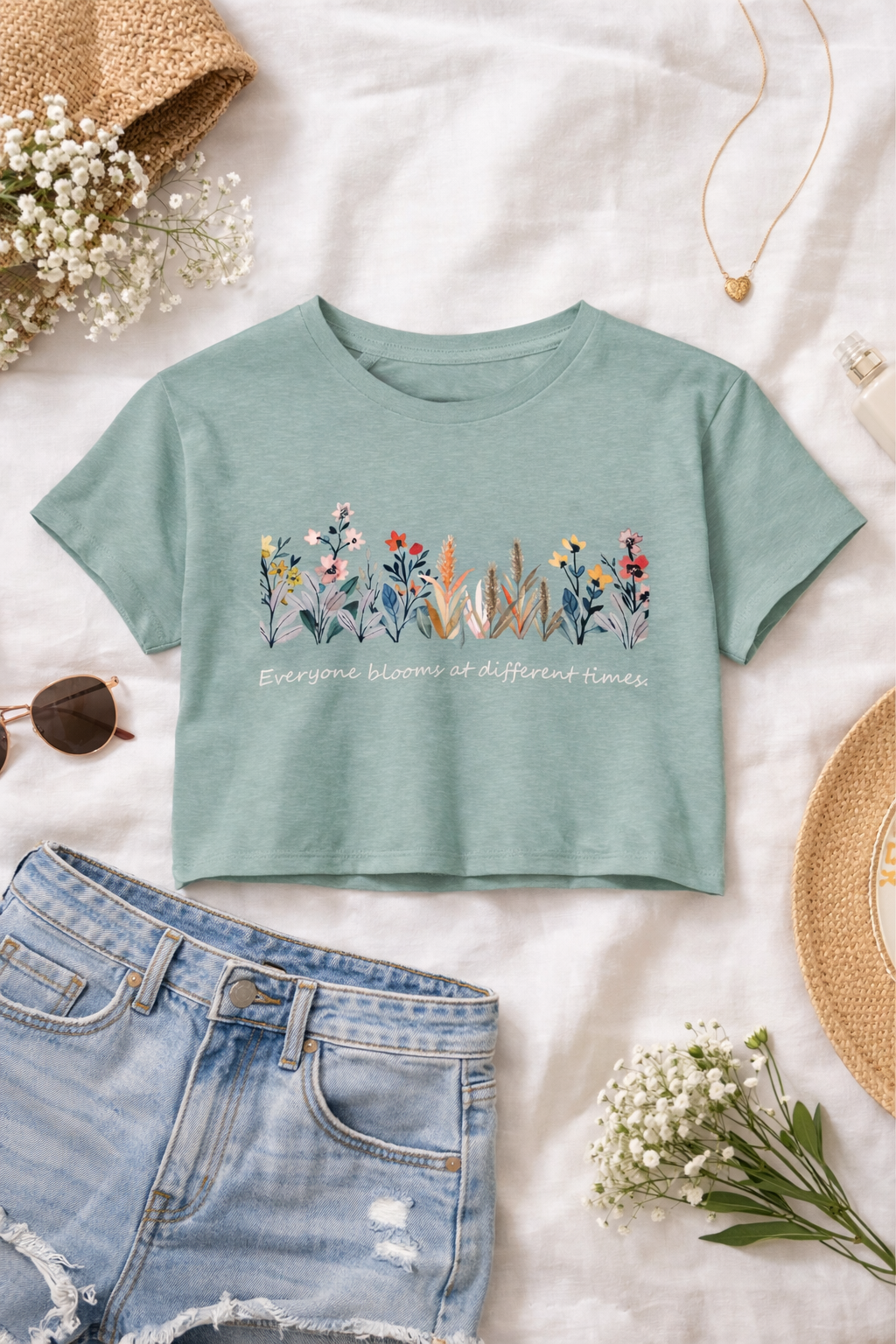 Mint green t-shirt with floral design and text on a white surface with denim shorts and flowers.